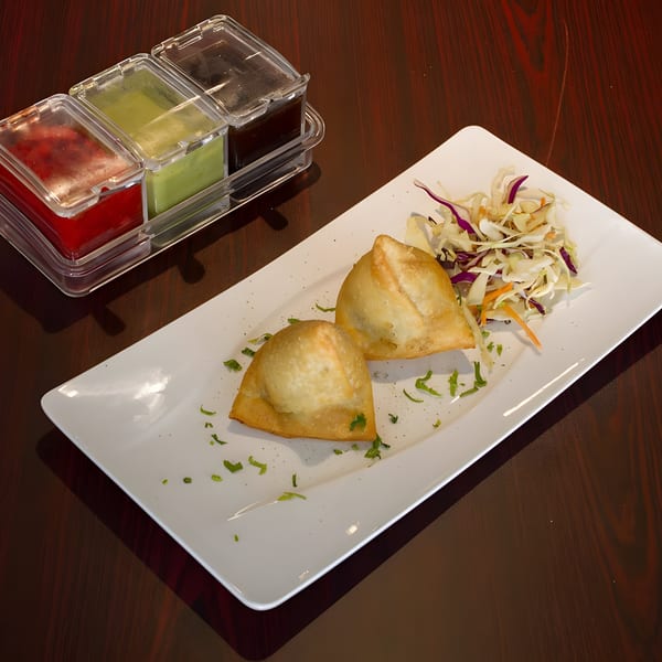 Two golden-brown samosas with a side of shredded cabbage and three dipping sauces on a wooden table.