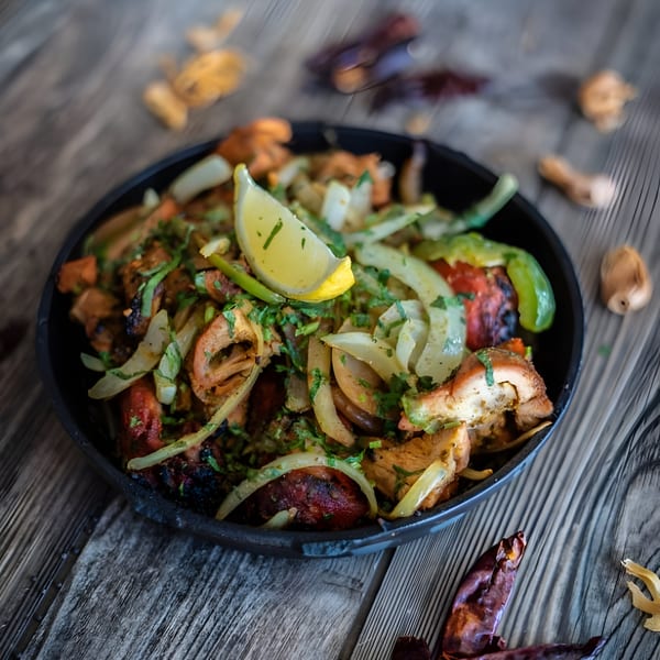 A close-up of Indian chicken tikka with onions, cilantro, and a lemon wedge in a black bowl.